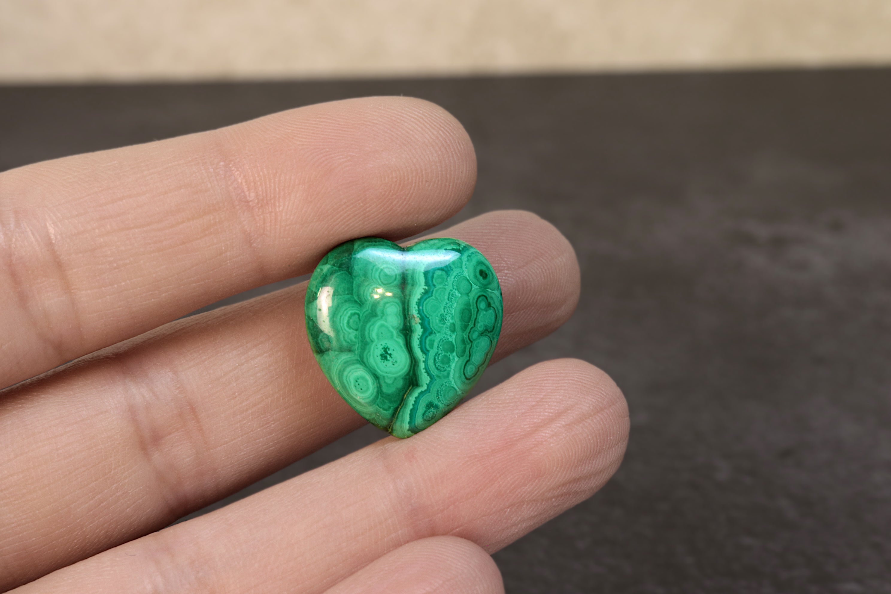 Green heart-shaped malachite stone held between fingers against a neutral background